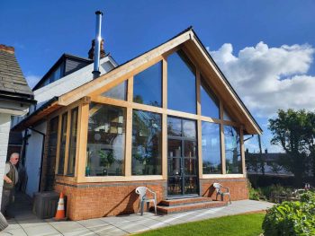 Exterior of a contemporary house extension with full-height glass windows and timber frame, featuring a brick base and patio area.